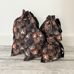 May include: A set of three drawstring bags in varying sizes, each featuring a repeating pattern of various cat faces in shades of brown, grey, orange, and black. The bags are constructed with black fabric drawstrings at the top for closure.