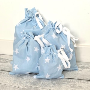 May include: A set of four light blue fabric drawstring bags of varying sizes, each featuring a repeating pattern of white five-pointed stars. Each bag is cinched at the top with a white ribbon tie, creating a gathered effect at the opening.