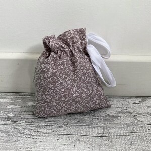 May include: A small, drawstring bag made of mauve fabric with a white floral pattern. The bag is tied with white ribbon. The bag is sitting on a distressed, grey wooden surface.