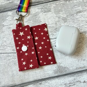 May include: A red fabric pouch featuring a white star pattern with a white snap closure, attached to a rainbow-striped lanyard with a silver metal swivel clasp, positioned next to a smooth, light grey rectangular wireless earbud charging case.