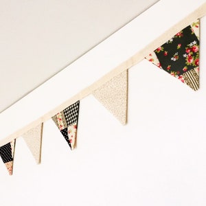 May include: A fabric bunting banner featuring a series of triangular pennants attached to a beige ribbon. The pennants alternate between a solid beige speckled pattern and a patchwork design containing black fabric with floral prints and black and white gingham.