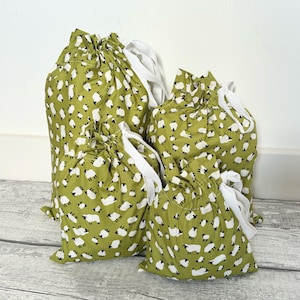 May include: A set of four drawstring bags in varying sizes, made from olive green fabric featuring a repeating pattern of small white sheep with black heads and legs. Each bag is cinched at the top with white fabric ribbons.