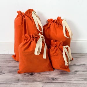 May include: A set of four orange fabric drawstring bags of varying sizes. Each bag is made from a solid orange textile and features a cream-coloured cotton ribbon drawstring closure at the top, gathered to secure the contents inside.