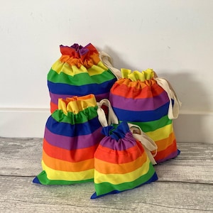 May include: A set of four drawstring pouches in varying sizes, each featuring horizontal stripes in red, orange, yellow, green, blue, and purple. The bags are made of fabric and include off-white cotton drawstrings to secure the top openings.