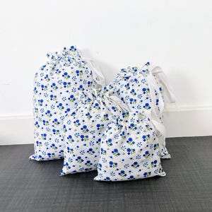 May include: Four white drawstring bags with a blue floral pattern. The bags are of varying sizes and are arranged together. Each bag has a white drawstring closure. The floral pattern includes blue flowers and green leaves.