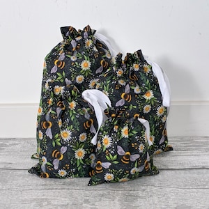 May include: A set of four drawstring bags in varying sizes, featuring a black fabric base with a repeating pattern of yellow and black bumblebees, white daisies, and green leaves. Each bag is cinched at the top with a white ribbon drawstring.