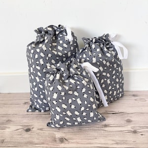 May include: Three grey drawstring bags with a white sheep pattern. The bags vary in size and have white ribbon closures. The bags are made of fabric and are on a wooden surface.