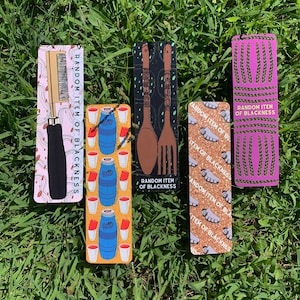 May include: Four colourful bookmarks with different designs. The bookmarks feature a comb, a pair of wooden spoons and a fork, a pattern of braided rope, and a pattern of red cups and blue barrels. Each bookmark has the text "Random Item of Blackness" printed on it.