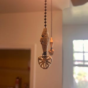 Può includere: Una catenella per ventilatore a soffitto con un cordino a perline marrone scuro attaccato a un ornamento color beige chiaro, avvolto e a forma di fuso con un motivo a spirale, che termina in una piccola gabbia sferica aperta in materiale chiaro.