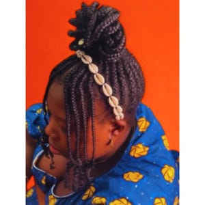 May include: A person with dark braided hair styled in a bun, decorated with a white shell headband. The person is wearing a blue top with yellow floral patterns, set against an orange background.