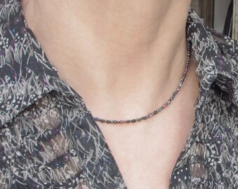 May include: A delicate necklace composed of tiny, multi-colored faceted beads in shades of dark gray, muted pink, and deep blue. The beads are strung in a repeating pattern, creating a subtle, textured appearance around the neck area.