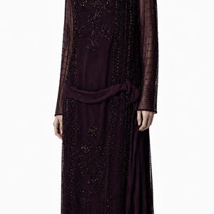 May include: A dark purple, long-sleeved dress featuring an overlay of sheer fabric adorned with intricate beadwork in a grid and floral pattern. The garment includes a dropped waistline with a side-tie detail and a straight, calf-length skirt.