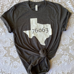 May include: A dark gray t-shirt with a white outline of the state of Texas and the number 76063 printed on it.