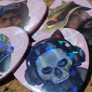 May include: Four heart-shaped badges featuring holographic finishes and illustrations of characters. One badge displays a skull mask, another a soldier in a cap with a flag, and others show figures with cat ears. The badges have a light pink background.