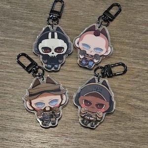 May include: Four acrylic keychains with black hardware. Each keychain features a cartoon character design. The characters have different skin tones and wear various outfits. The keychains are on a wooden surface.