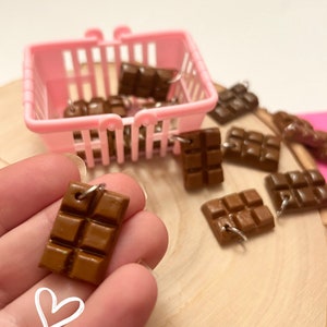 Op de afbeelding: Een roze plastic winkelmandje gevuld met kleine chocolade reep charms. De charms zijn bruin en hebben een zilveren lus om op te hangen. De charms liggen verspreid op een houten oppervlak.