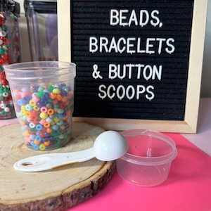 May include: A clear plastic cup filled with colourful beads, a white scoop, and a small clear container sit on a wooden slice. A black felt board with the words "BEADS, BRACELETS & BUTTON SCOOPS" is in the background. The background is pink and purple.