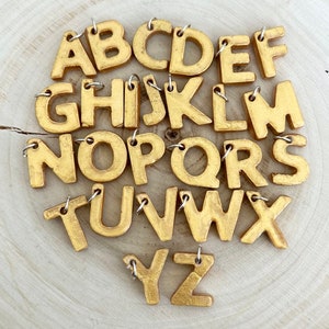 Può includere: Una collezione di ciondoli dorati a forma di lettere dell'alfabeto, ciascuno con un piccolo anello color argento per l'attacco. Le lettere sono disposte in un ordine apparentemente casuale, con tutte le 26 lettere dell'alfabeto visibili. I ciondoli sono su una superficie di legno chiaro.