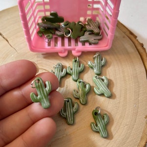 Può includere: Una collezione di ciondoli a forma di cactus verdi e bianchi con anelli di salto argentati. I ciondoli sono in un cestino di filo rosa e su una superficie di legno.
