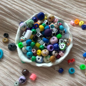 Könnte beinhalten: Eine weiße Schüssel gefüllt mit bunten Plastikperlen. Die Perlen haben verschiedene Formen, darunter Buchstaben, Sterne und Kreise. Einige Perlen sind durchsichtig, andere undurchsichtig. Die Perlen sind in einem zufälligen Muster angeordnet.