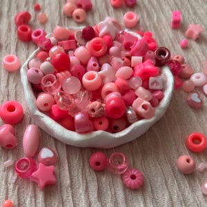 May include: A selection of pink and red beads in diverse forms, such as round, star, and heart shapes, overflowing from a white bowl. The beads are spread across a textured, light-coloured surface.
