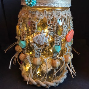 May include: A glass jar decorated with twine, beads, and fabric fish. The jar has a light brown lid with a blue flower on top. The jar is lit from within with white fairy lights.