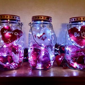 May include: Three glass mason jars with red lids and wire handles, each filled with fairy lights and red heart decorations. One jar has "xoxo" on a heart, another has a heart, and the third says "Be Mine".