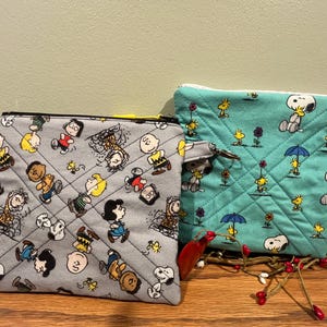 Op de afbeelding: Twee gewatteerde stoffen etuis met Peanuts-personages. Eén etui is grijs met een zwarte rits en een patroon van Charlie Brown, Snoopy en andere personages. De andere is turquoise met een gele rits en een Snoopy- en Woodstock-patroon.