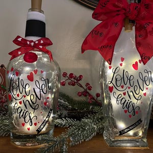 May include: Two clear glass bottles decorated with red hearts and the words "Love never fails" in black script. Each bottle has a red bow and string lights inside. The bottles are part of a home decor theme.