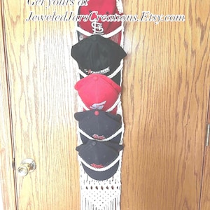 May include: A macrame cap or hat holder with multiple baseball caps in red and black. The holder is white with a fringe at the bottom and a metal ring at the top for hanging. Text on the holder reads "JeweledJarsCreations.Etsy.com".