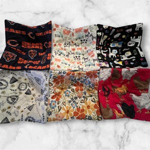 Pode incluir: Uma coleção de protetores de tigela de tecido com vários padrões. Os designs incluem logotipos do Chicago Bears, animais de fazenda, gatos com texto "Meow", estampas florais e galinhas. Os protetores são quadrados com cantos dobrados.