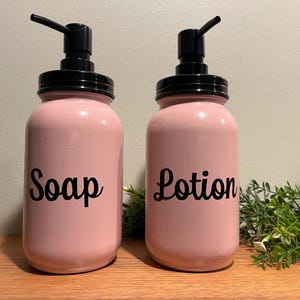 May include: Two pink glass pump bottles with black pumps and lids. The bottle on the left says "Soap" and the bottle on the right says "Lotion".