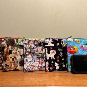 May include: Six colourful zippered pouches with various animal prints. The pouches are lined up on a wooden surface. The pouches feature a variety of animal prints, including dogs, cats, and fish. The pouches are all different sizes and shapes.