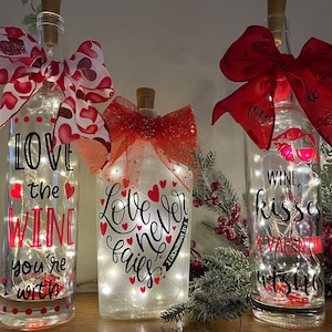 May include: Three decorative wine bottles with fairy lights inside. Each bottle has a different message in black script: "Love the wine you're with", "Love never fails", and "Wine, kisses & Valentine wishes". Red bows and heart accents adorn the bottles.