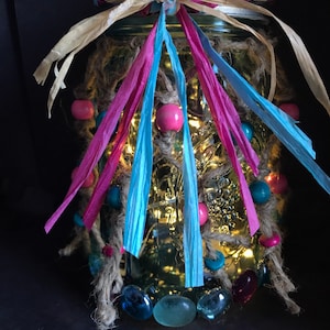 May include: A glass jar decorated with pink, blue, and brown ribbons, beads, and glass stones. The jar is lit from within with white fairy lights.