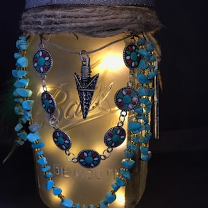 May include: A mason jar with a gray lid and a brown twine bow. The jar is decorated with turquoise and red beaded necklaces and a silver arrow pendant. The jar is lit from within with white fairy lights.