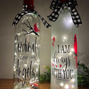 May include: Two decorative glass bottles with fairy lights inside. One bottle has the text "Cardinals appear when Angels are near" and a red cardinal design. The other bottle has the text "I am always with you" and a red cardinal design. Both bottles have a black and white checkered ribbon.