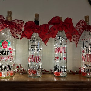May include: Four decorative wine bottles with cork stoppers and red heart-patterned bows. Each bottle has a different message in black and red, with fairy lights inside. Messages include "Best Friends Forever" and "Love the Wine."