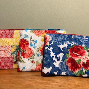 May include: Three quilted zippered pouches with floral patterns. The first pouch is a patchwork design with red, yellow, and green squares. The second pouch has a white background with red and blue flowers. The third pouch has a white background with blue and red flowers.
