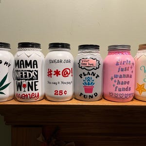 Puede incluir: Seis tarros de vidrio pintados de blanco con diferentes lemas y diseños. Los tarros están etiquetados con "Go Fund My", "Mama Needs Wine Money", "Swear Jar #*@! You say it. You pay it. 25¢", "Your name here. Plant Fund", "Girls Just Wanna Have Funds" y "Vacation Fund".