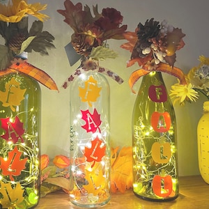 May include: Three glass bottles illuminated with internal string lights, each decorated with autumn-themed leaf cutouts spelling FALL. The bottles are topped with artificial sunflowers, pinecones, and foliage, accented by plaid ribbons.