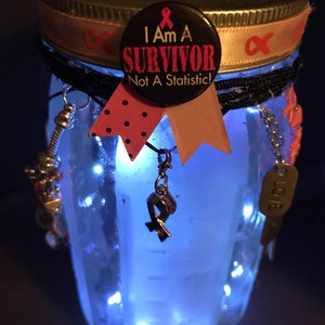 May include: A glass jar with a pink ribbon and a button that says "I am a Survivor Not a Statistic!" with a pink ribbon on it. The jar is filled with lights and other decorations.