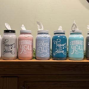 May include: A collection of six decorative tissue holders made from painted mason jars. Each jar is a different color, including white, pink, blue, and teal, and features a decorative lace trim around the lid. The jars have phrases like "Here's a tissue for your issue" and "Bless You."