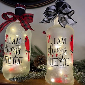 May include: Two frosted glass bottles illuminated with internal string lights. Each bottle features a red cardinal illustration, small red hearts, and the black text I AM always WITH YOU. One bottle has a red plaid bow, the other a black and white buffalo check bow.