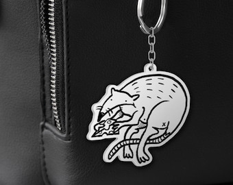 May include: A silver-coloured keyring with a raccoon design. The raccoon is illustrated in black and white, holding a small object. The keyring is attached to a black backpack.