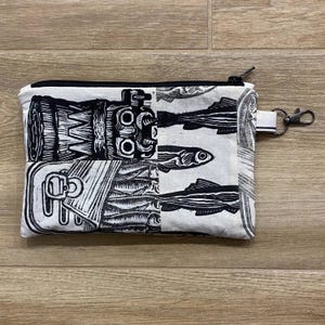 Könnte beinhalten: Eine weiße Reißverschlusstasche mit einem Schwarz-Weiß-Druck. Der Druck zeigt eine Kombination aus abstrakten Formen und Fischillustrationen. Die Tasche hat eine kleine Schlaufe mit einem Metallverschluss.