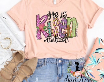 May include: A pink t-shirt with the words "He is Risen indeed!" in colorful patchwork letters. The letters are decorated with flowers, crosses, and other Easter-themed designs.