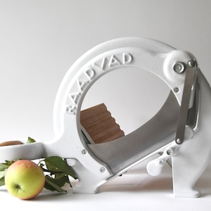 Puede incluir: Cortador de manzanas de hierro fundido blanco con una tabla de cortar de madera y una manzana verde con hojas. El cortador tiene una palanca y un mango. El nombre de la marca "RAADVAAD" está grabado en el lateral.