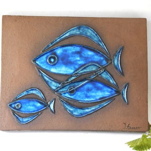 May include: A rectangular brown ceramic wall plaque featuring three raised, glossy blue fish with dark outlines. The fish are arranged in a school, with two larger fish overlapping and one smaller fish to the left. The signature J. Exner is visible.