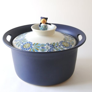 Puede incluir: Una cacerola de cerámica azul con una tapa blanca que presenta un diseño floral en azul, verde y amarillo. Una pequeña muñeca de madera con un vestido azul se sienta en la parte superior de la tapa.